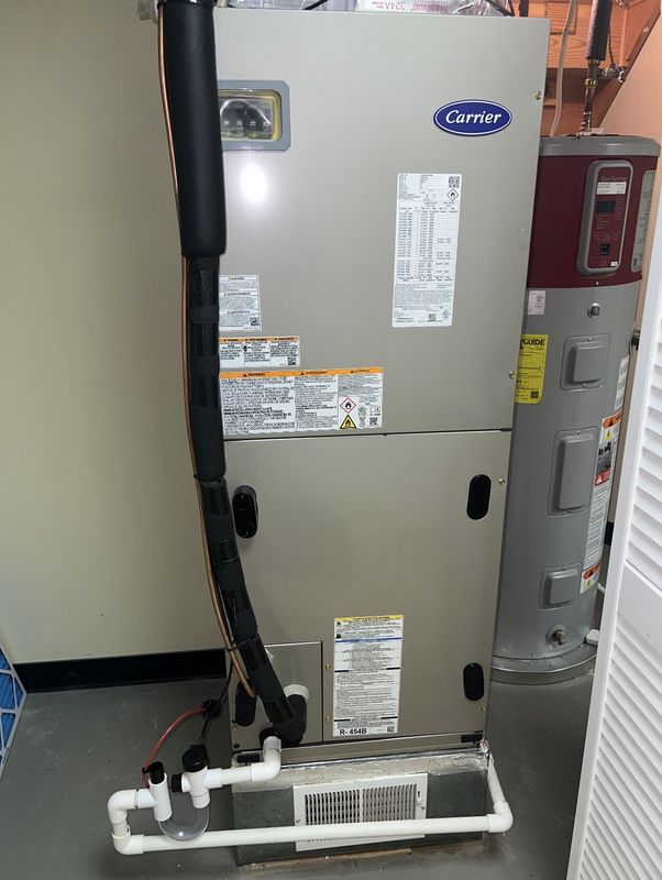 A new Carrier air handler in a utility closet, labeled for R-454B refrigerant, with the insulated refrigerant line down the front and a PVC condensate trap at the base.
