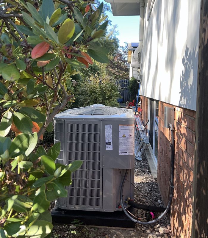 A new outdoor unit on a pad in a narrow side yard between shrubs and a brick house, with the refrigerant lines and electrical connected at the corner.