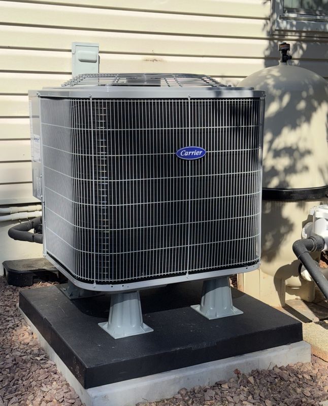 A new Carrier outdoor unit raised on riser feet on a composite pad beside a house with vinyl siding.